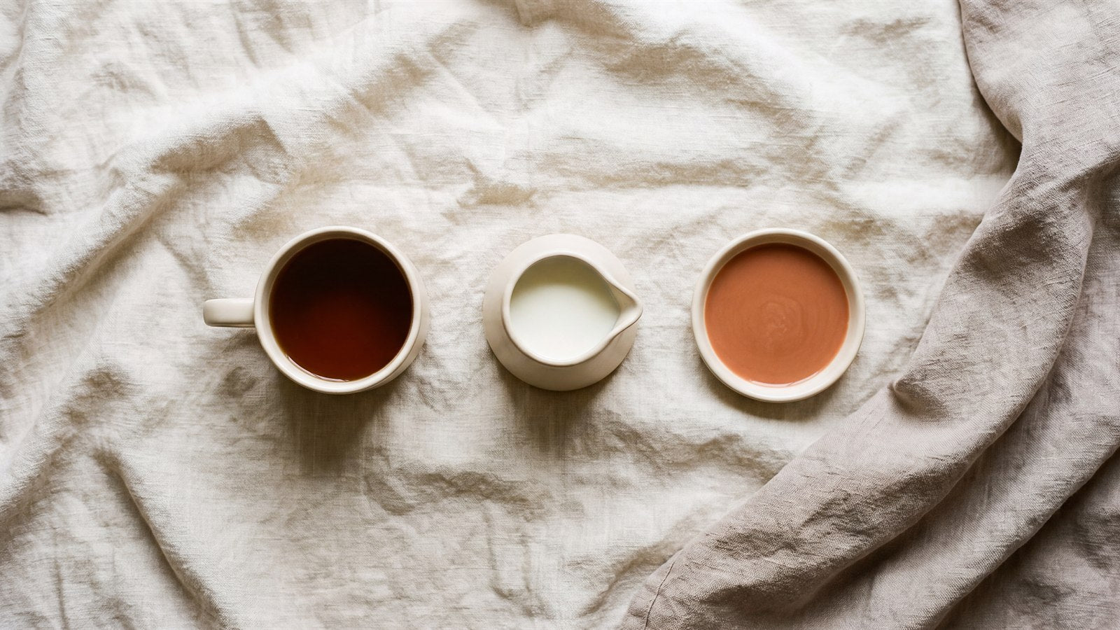A cup of dark tea, a small jug of milk and a dish of terracotta liquid in a row on linen