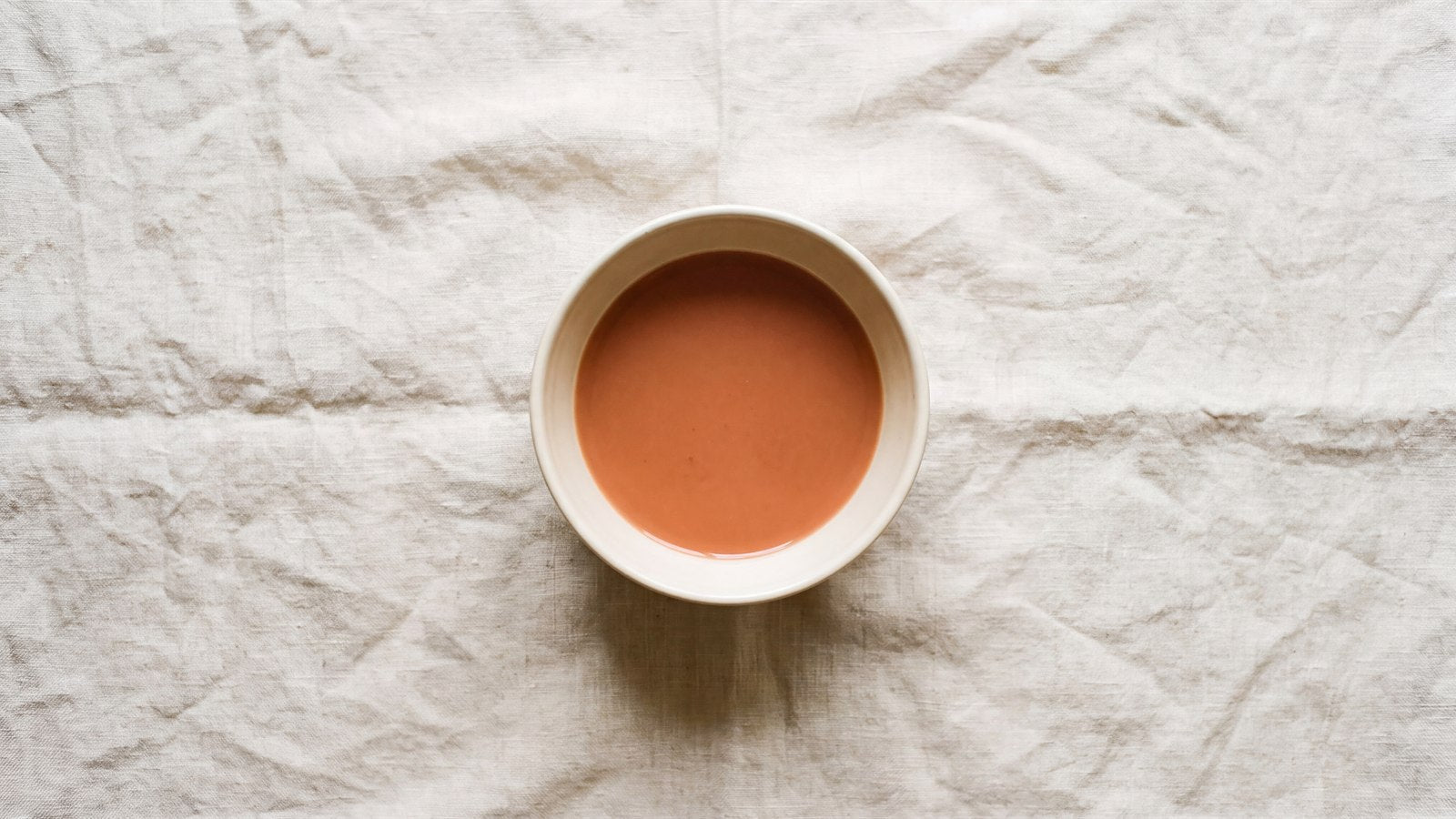 A single ceramic bowl of terracotta liquid, still and centred on a wide field of linen