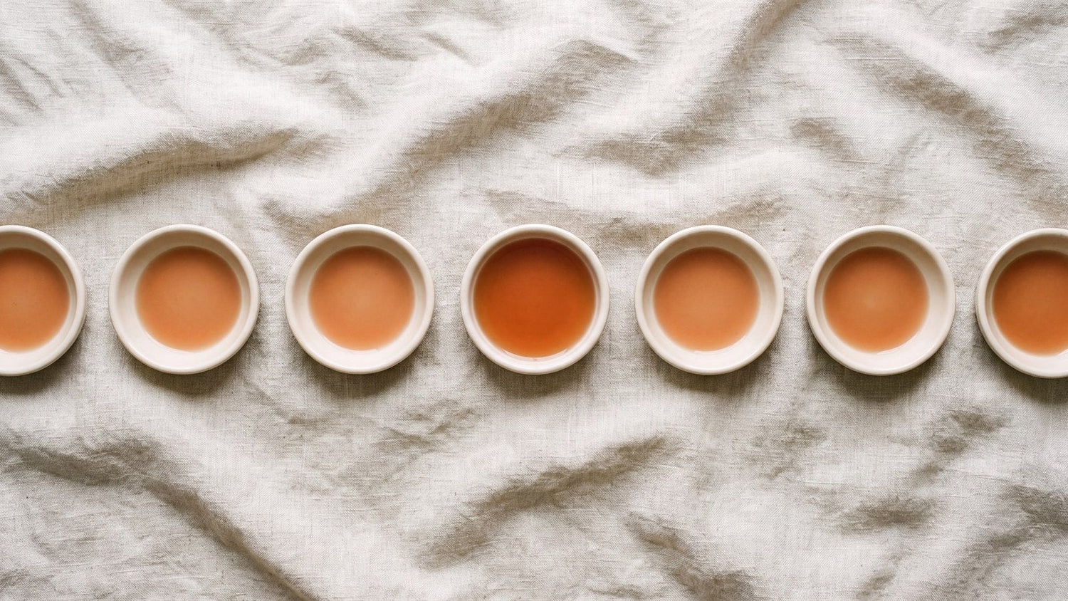 A row of identical ceramic dishes holding a shallow film of terracotta liquid, one holding noticeably more