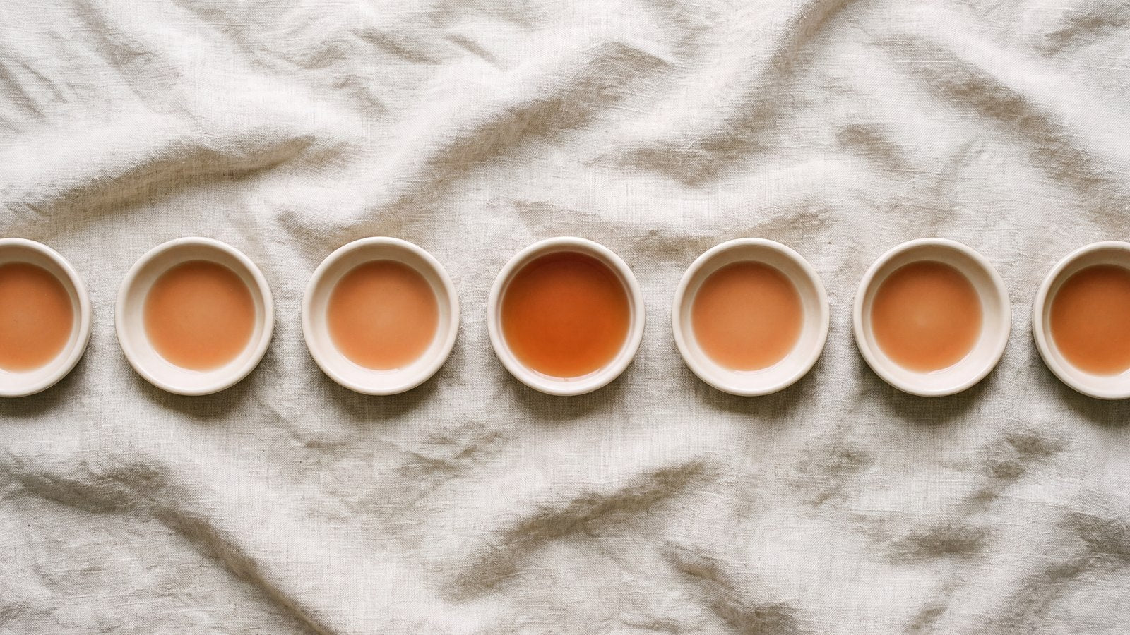 A row of identical ceramic dishes holding a shallow film of terracotta liquid, one holding noticeably more