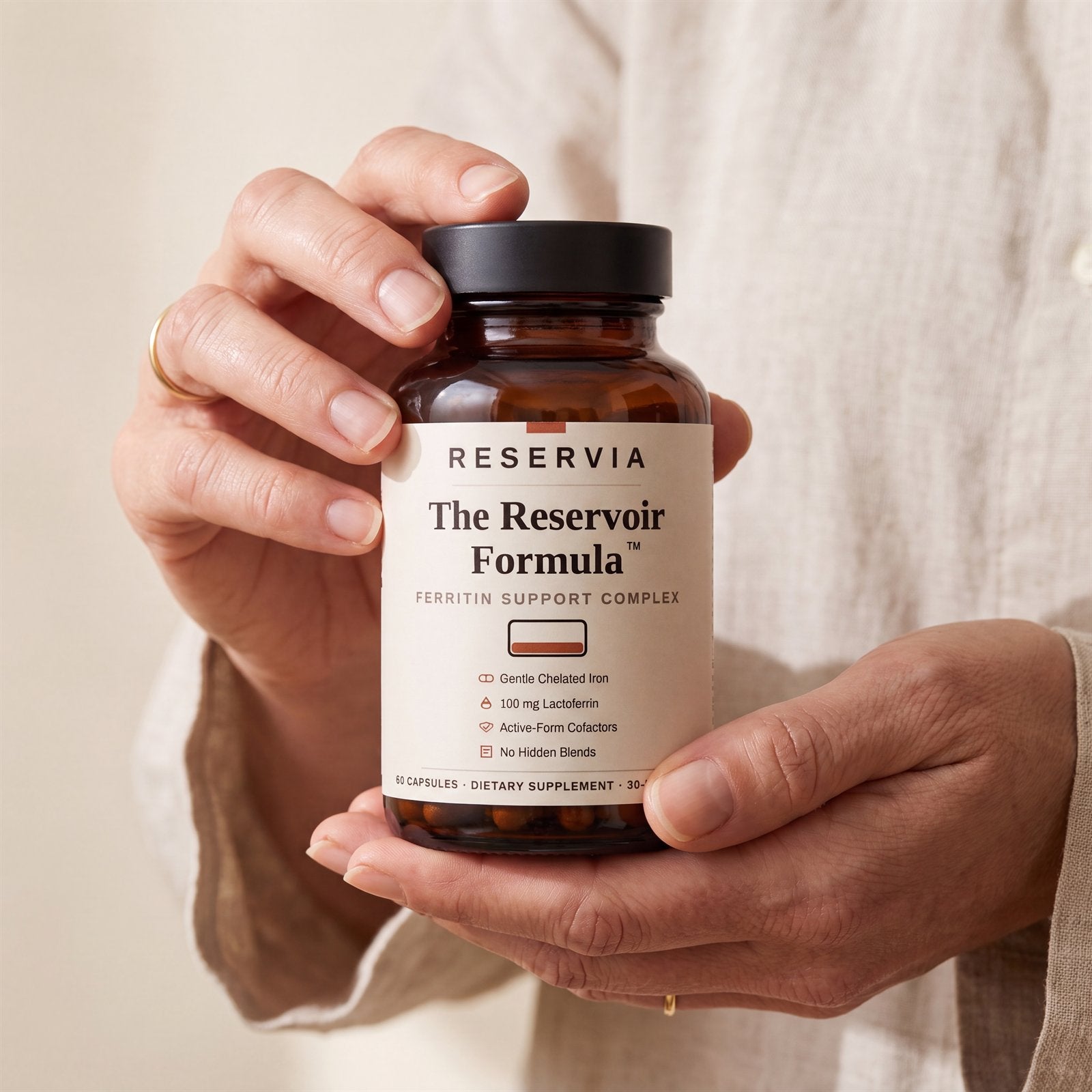 Two hands holding The Reservoir Formula bottle, the front label fully readable: gentle chelated iron, 100 mg lactoferrin, active-form cofactors, no hidden blends.