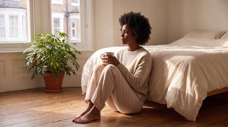 A woman sitting on a sunlit bedroom floor with her back against the bed, holding a mug